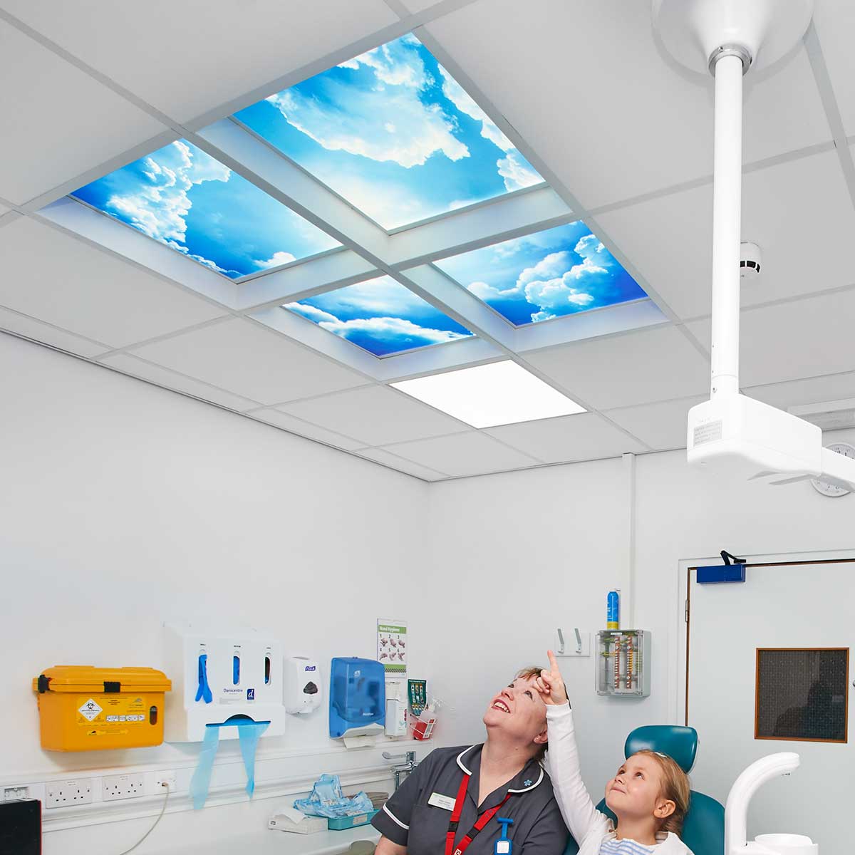Nurse & little girl pointing & looking at LED ceiling lights which display a vivid bright blue sky with white clouds.